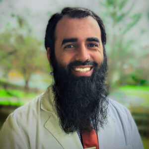 Headshot Of Doctor Mohammad Khubaib, MD, CAQSM From The Shoulders Up Smiling In Front Of A Nature Background.